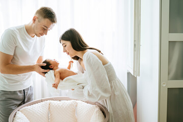 Young husband with beautiful wife holding newborn baby girl standing near crib happy family parenthood concept modern home bedroom interior.