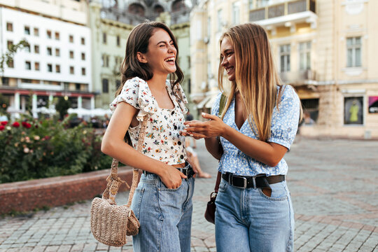Brunette and blonde happy girls in trendy outfits laugh and walk outside. Attractive women in denim pants and stylish floral blouses smile and talk.