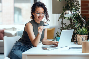 Cute woman holding white credit card for shopping online with computer while sitting in living room...