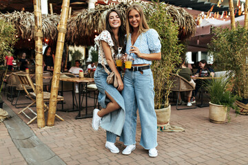 Obraz na płótnie Canvas Cheerful curly brunette woman in trendy denim pants and white blouse walks with beautiful friend. Attractive blonde lady holds glass of lemonade.