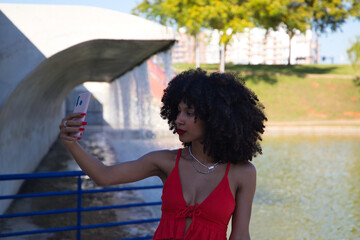 Obraz premium beautiful afro-american woman taking a selfi. The woman is wearing a red top and looks attentively at the camera of the phone to look perfect and beautiful. life style concept