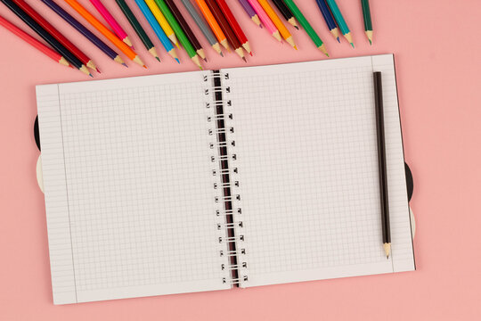 Open school squared notebook and colorful pencils on the pink background. Blank white sheet of paper book on the table. Office supplies on the desktop. Back to school. Copy space for text. Top view