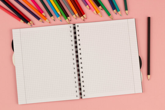 Open school squared notebook and colorful pencils on the pink background. Blank white sheet of paper book on the table. Office supplies on the desktop. Back to school. Copy space for text. Top view