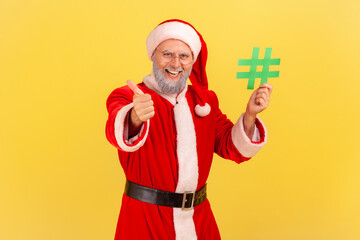 Fototapeta na wymiar Smiling elderly man with gray beard wearing santa claus costume holding green hashtag in hands and showing thumb up, recommended website. Indoor studio shot isolated on yellow background.
