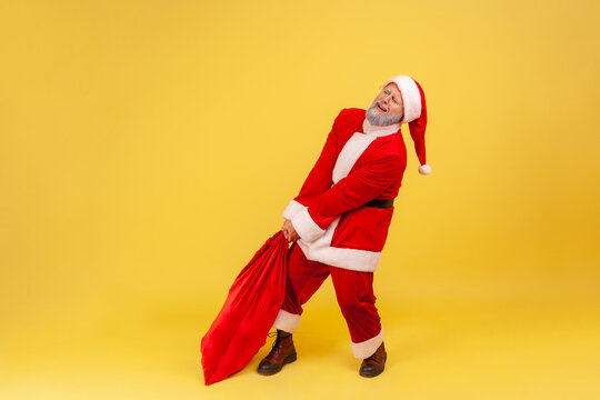 Elderly Man With Gray Beard Wearing Santa Claus Costume Dragging His Very Large And Heavy Bag Full Of Presents, Frowning Face, Trying To Lift Up. Indoor Studio Shot Isolated On Yellow Background.