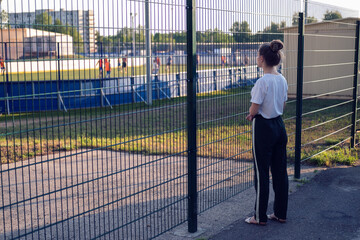 A girl stands and looks through the fence at the athletes playing at the stadium. The girl wants,...