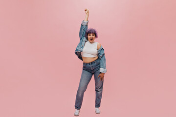 Charming woman in good mood with short hair in tight jeans and wide blue jacket shows her tongue at camera on pink background..