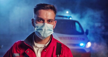 Portrait Caucasian handsome young man paramedic in medical mask. Attractive male doctor in respirator looking at camera. Tired physician at night during covid-19 pandemic. Ambulance car on background.