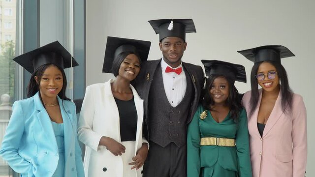 A young cheerful young man and four happy girls, university graduates stand in front of the camera in master's hats. Sincere smile