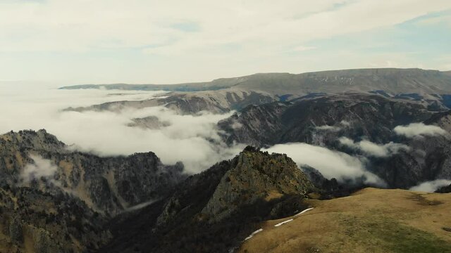 Aerial view of Caucasus Mountains. Gray sky is visible over green meadows. White clouds form river. Peaks of sharp mountains are covered with glaciers and snow in the distance. Cloudy weather