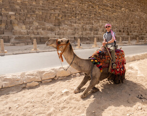 A tourist on a camel poses against the backdrop of the pyramids in Giza, Cairo, Egypt. Kufiya is wrapped around the man's head. Camel knelt down.