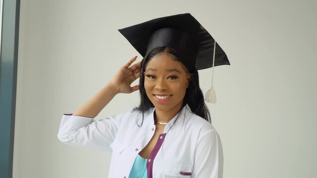 A young African American female graduate in a master's hat and white medical gown smiles poses for the camera. An important event. Young specialist