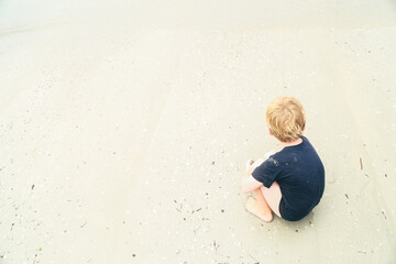 Fototapeta premium Little boy sitting on beach surrounded by smooth sand with copy space