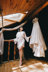Fototapeta na wymiar A beautiful brunette in lingerie stands in front of a hanging long white dress with a long veil in a wooden hotel room. Wedding photography.