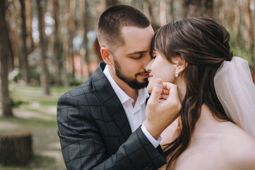 Wedding portrait of newlyweds in love close-up. A bearded, stylish groom in a suit straightens his hair, the hairstyle of a beautiful bride in a white dress in nature.