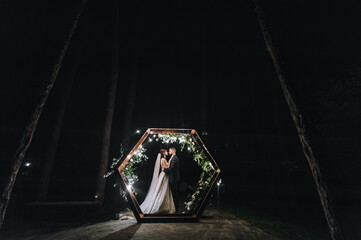 Stylish groom in a vest and a cute brunette bride in a white long dress with a veil tenderly hug...