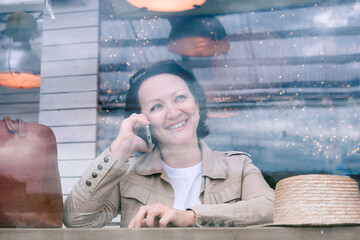 A smiling woman is sitting at a table on the summer terrace of a cafe and talking on the phone.