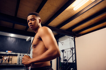 African American male looking serious into the camera. Topless mixed race male exercising indoors in the gym with skipping rope. High quality photo