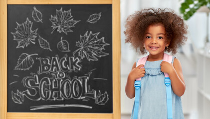 childhood and education concept - happy little african american girl with backpack over chalkboard...
