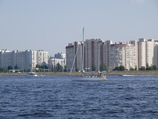 A white sports yacht with white sails in the bay against the background of multi-storey buildings. Training of yachtsmen on a sunny summer day.