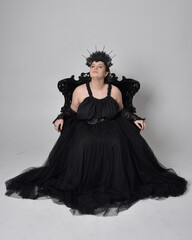 Fototapeta premium Full length portrait of young plus sized woman with short hair, wearing long black tulle gothic gown with crown and gloves, sitting pose on a ornate throne chair on light studio background.