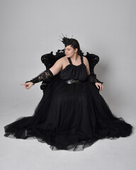 Full length portrait of young  plus sized woman with short  hair,  wearing long black tulle gothic gown with crown and gloves, sitting pose on a ornate throne chair on light studio background.