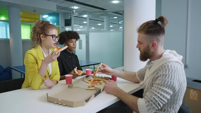 Cheerful young people in relax zone of modern work space, eating pizza and joking. Arc shot multiethnic pals having lunch break. Concept of fast food