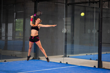Woman playing padel in a blue grass padel court indoor - Young sporty woman padel player hitting ball with a racket
