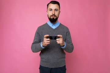 handsome good looking brunet bearded young man wearing grey sweater and blue shirt isolated on pink background with empty space holding in hand and using mobile phone communicating online looking at