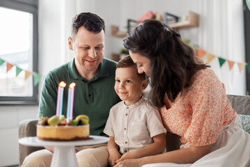 family, holidays and people concept - portrait of happy mother, father and little son with four candles on birthday cake sitting on sofa at home party