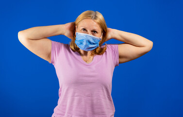 Middle aged woman with quirurgical mask stretching with hands on head on blue studio background.