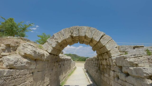 Olympia, Greece POV at Archaeological site area that hosted ancient sports Games. Walking to the stone built arch and entrance of the stadium of athletic contests on classical times, against blue sky.