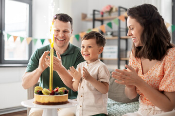 family, holidays and people concept - portrait of happy mother, father and little son with firework candle burning on birthday cake sitting on sofa at home party