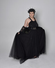 Naklejka premium Full length portrait of young plus sized woman with short hair, wearing long black tulle gothic gown with crown and gloves, standing pose on light studio background.