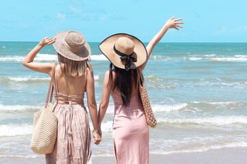 Two beautiful Asian women with big hat from behind enjoy spending time on tropical sand beach blue sea together, resting and relaxing on summer holiday vacation.