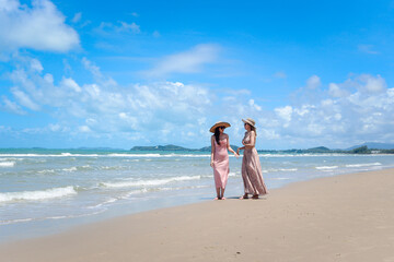 Two beautiful Asian women with big hat and sunglasses enjoy spending time with friend on tropical sand beach blue sea together, go for a walk, resting and relaxing on summer holiday vacation.