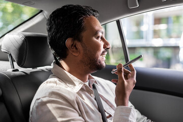 transport, business and technology concept - happy smiling indian male passenger or businessman using voice command recorder on smartphone on back seat of taxi car