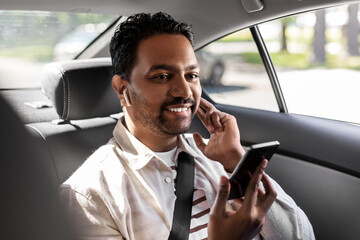 transport, business and technology concept - happy smiling indian male passenger with wireless earphones using smartphone on back seat of taxi car