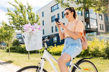 people, leisure and lifestyle - happy young woman with smartphone, backpack and flowers in basket of bicycle on city street