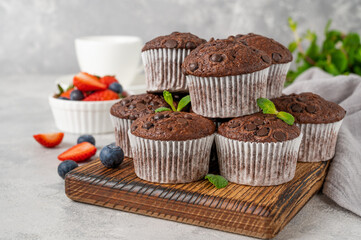 Chocolate muffins or cupcakes with chocolate drops on a wooden board on a gray background with fresh berries and mint. Copy space.