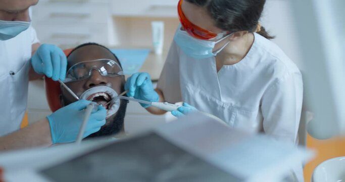  Close-up an African American male patient in optragate is sitting with open mouth and having teeth cleaning procedure by using a dental brush, mirror and female assistant is using a saliva ejector.