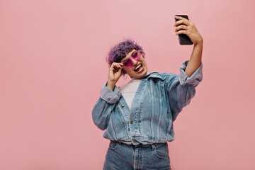 Naughty girl with short lilac hair in pink glasses laughs on isolated background. Fashionable woman holding smartphone and posing for camera..