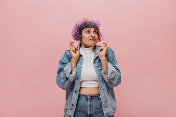 Cute adult girl in denim suit and short light top posing on isolated background. Woman with purple haircut crosses her fingers..