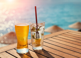 Glass of cold beer and soda lemon with ice on wooden table in a hot  summer day. Summer drink with blurry beach on background. Sunny day on vacation.
