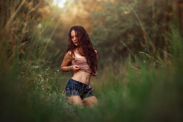 Sexy woman posing in a field in the grass rays of the sunset. Lifestyle on nature dressed in jeans shorts and beige top in golden hour. Romantic autumn mood, long tall grass in the meadow