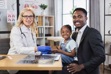 Qualified pediatrician monitoring saturation of cute african girl using digital pulse oximeter. African man and her daughter visiting caucasian doctor. People smiling in camera.