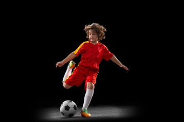 One little male football soccer player, boy training with football ball isolated on dark studio...