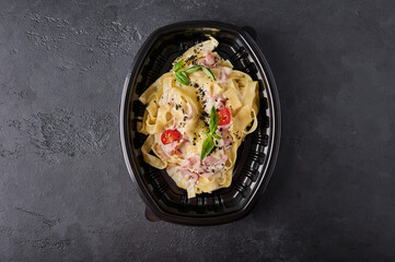 Tagliatelle carbonara in black plastic container on dark graphite background top view. Designer food concept for take away or delivery