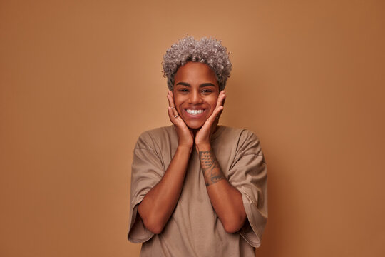 Portrait of cute young dark skinned woman with blonde hair and gentle smile holding hands under chin looking contented and carefree. Pretty biracial female student posing against brown background