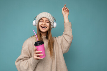 Beautiful happy smiling young blonde woman wearing beige winter sweater and hat isolated over blue background drinking beverage wearing white wireless bluetooth headphones listening to cool music